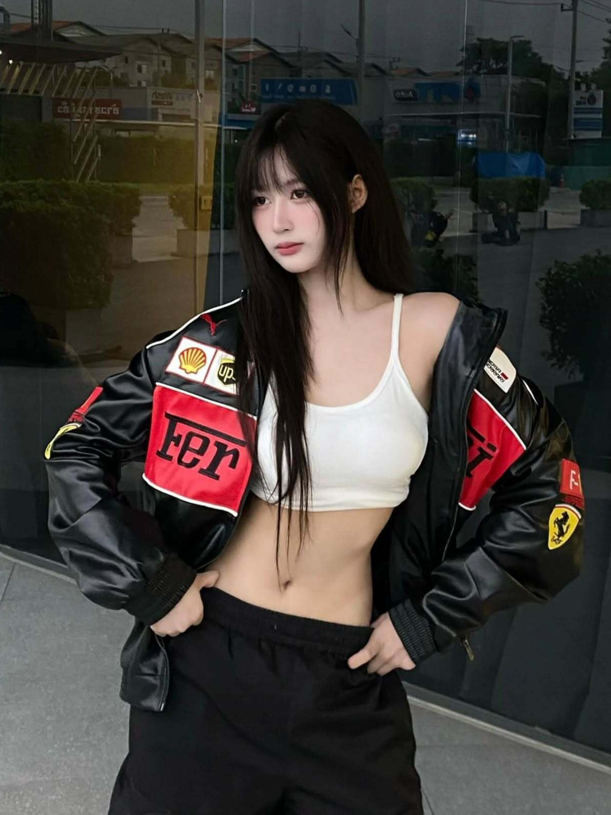 Person wearing a black jacket with logos and a white top, standing in front of a reflective surface.